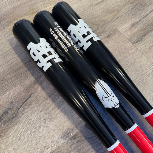 Three custom Caliburn fungo bats with black barrels, red handles, white team logos, and a white Caliburn sword logo, arranged on a wood surface.