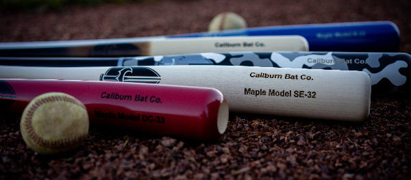Four Callburn Bat Co. maple baseball bats in different colors (blue, white camo, white, and red) arranged on a dirt field with baseballs in the foreground.