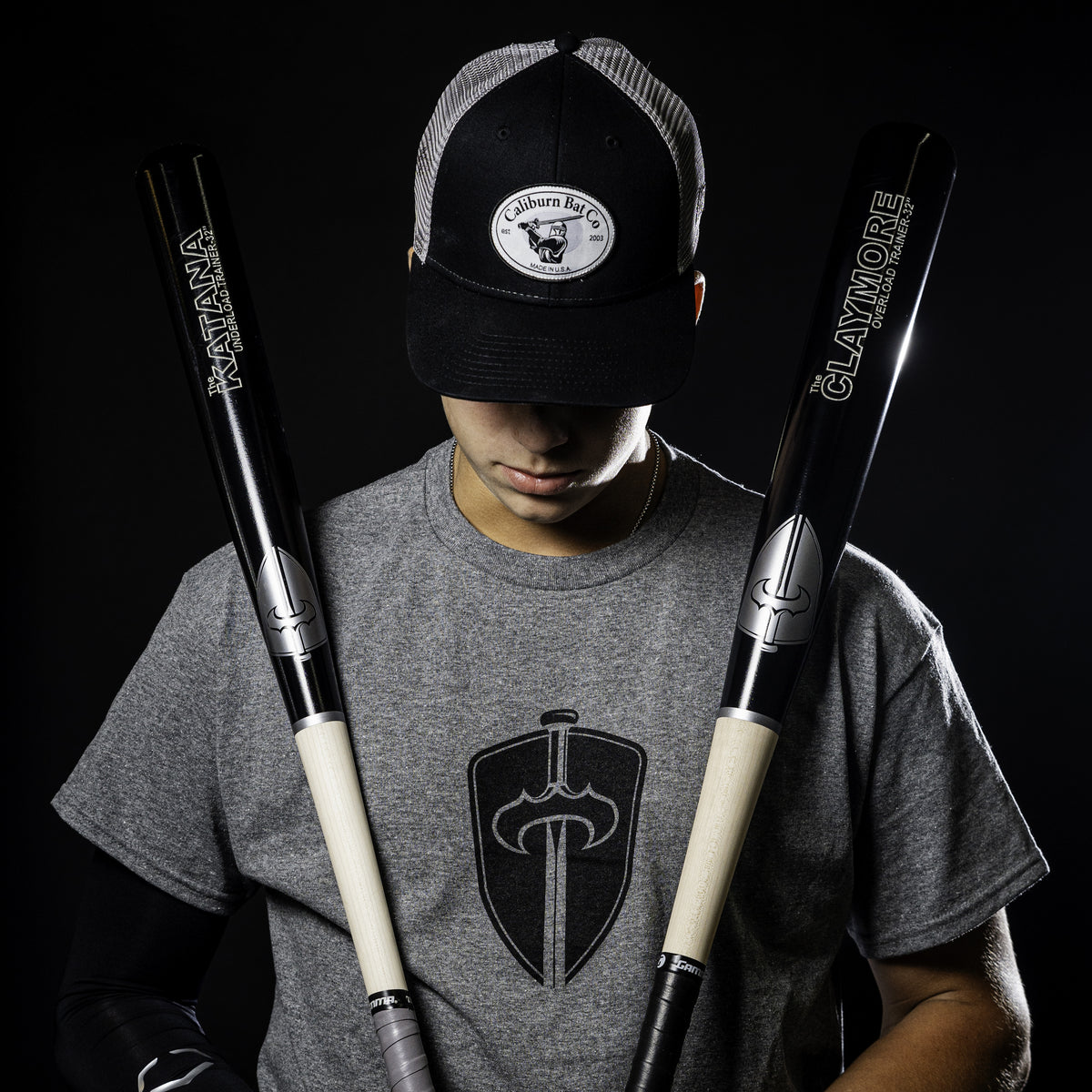 Player wearing a Caliburn cap and shirt holding Katana underload and Claymore overload training bats with black barrels and natural wood handles.