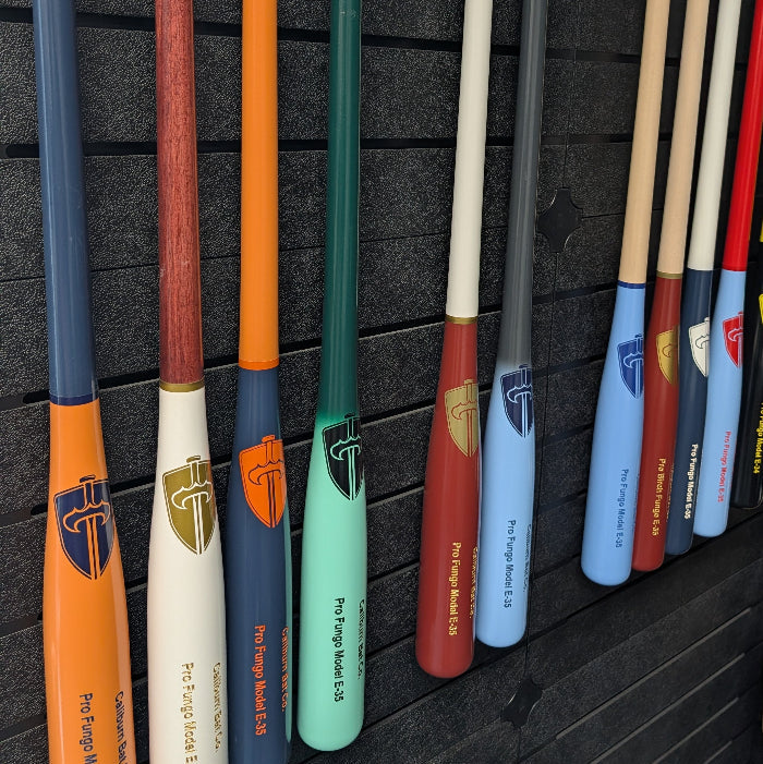 Caliburn fungo bats hanging on a black display wall, showing custom barrel and handle color combinations with Caliburn sword logos.
