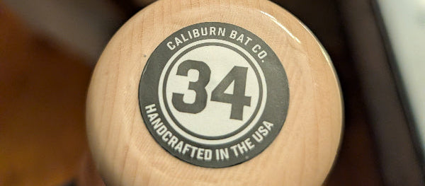 Close-up of the top of a wooden baseball bat showing a circular Caliburn Bat Co. stamp with the number 34, held in a hand with a blurred indoor background.