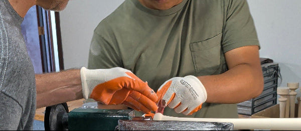 Two men wearing protective gloves and glasses work at a woodworking table, with one using a joint sanding attachment on a piece of wood. A gray dust collection hose is in the foreground, and a workshop environment is visible in the background.