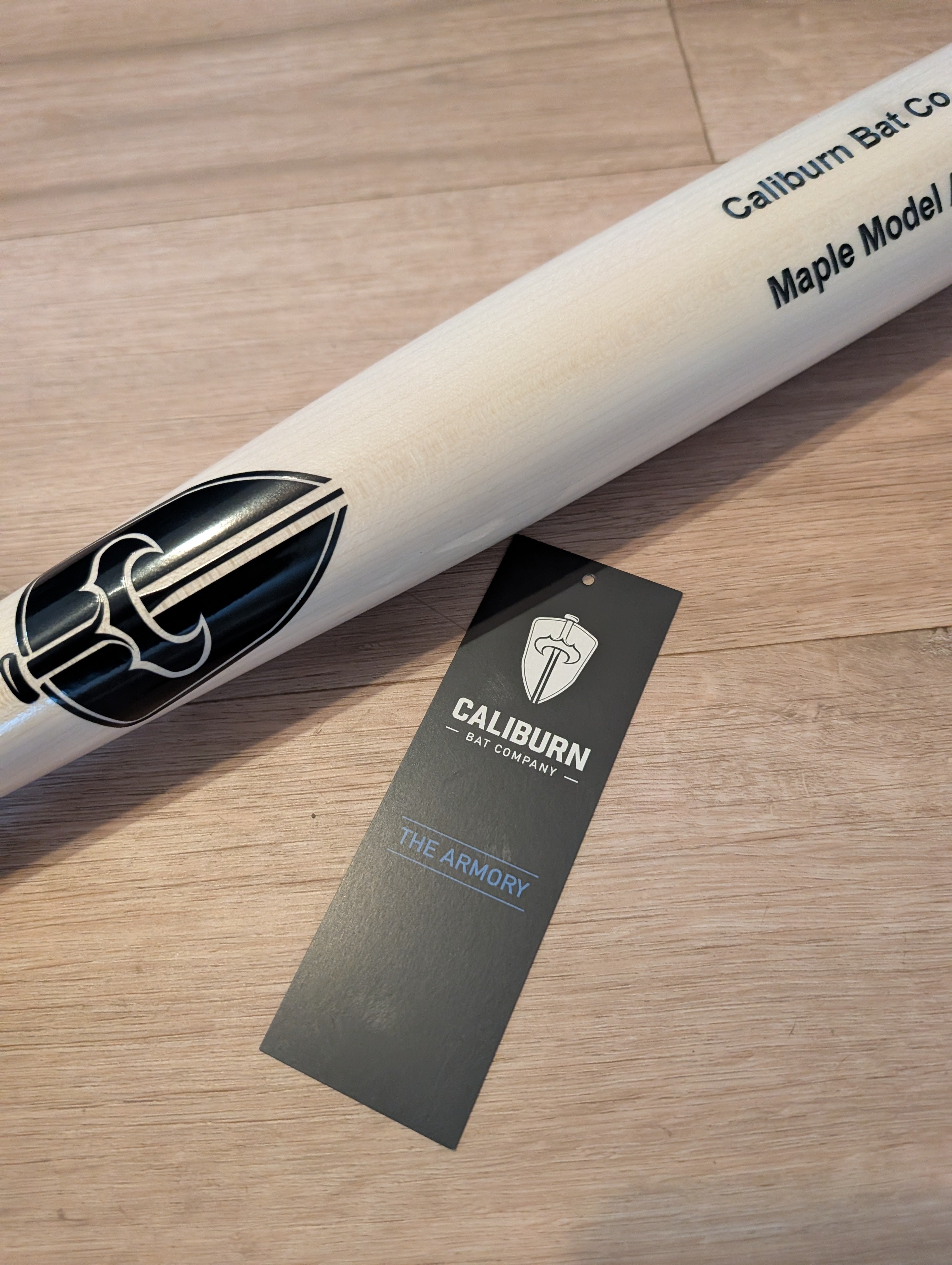 Caliburn maple bat with an Armory hang tag on a wood floor